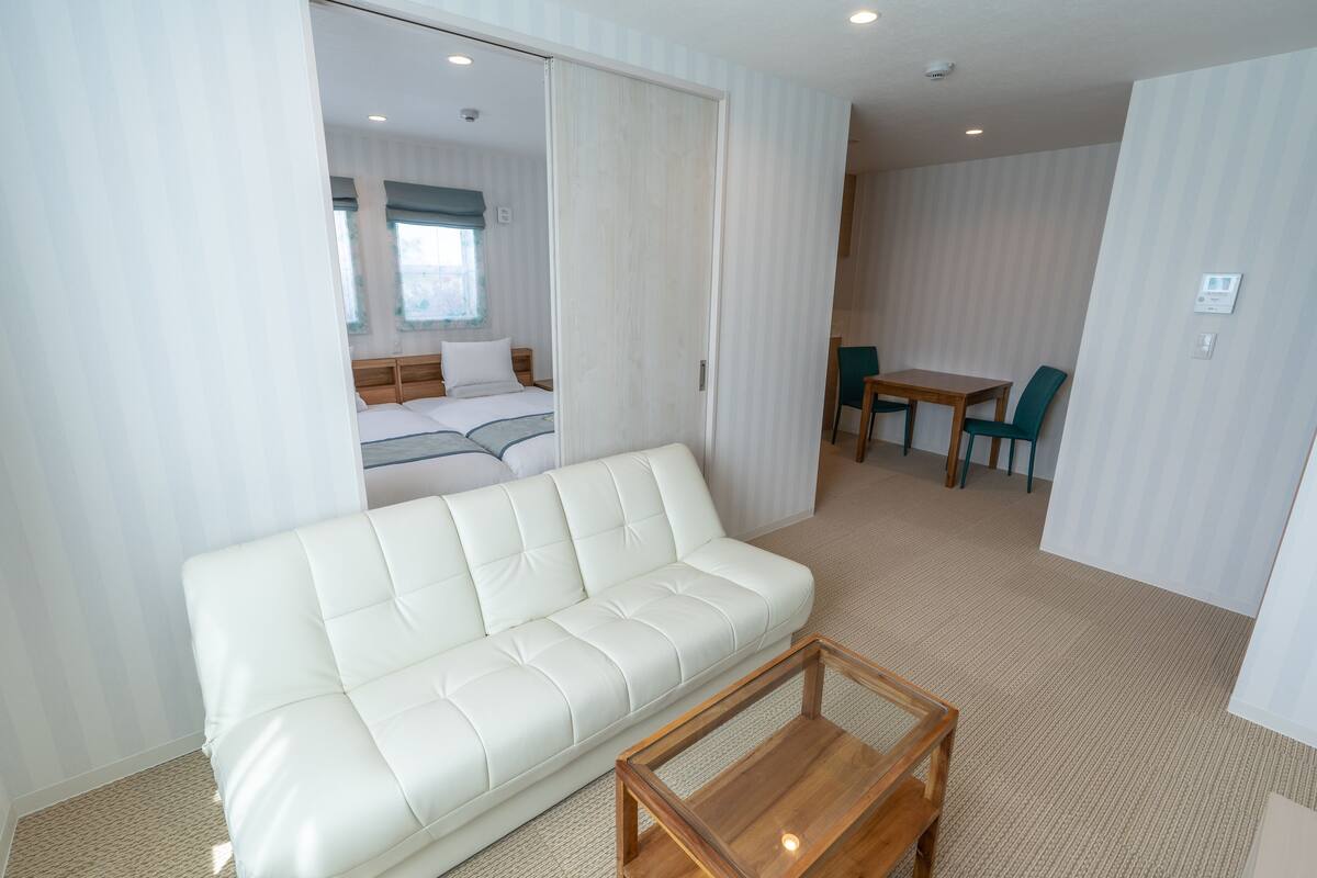 A modern hotel room with a white leather sofa, a wooden coffee table, a dining area with a table and chairs, and a sliding door leading to a bedroom.