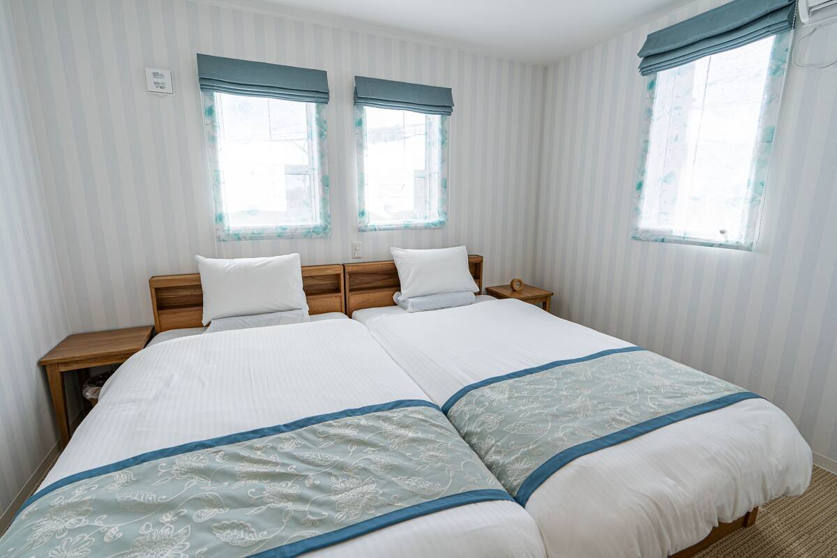 A double bed with a white and blue bedspread, two bedside tables, and two windows with blue curtains.