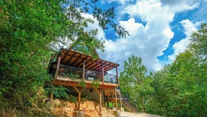A wooden cabin with a balcony, surrounded by lush greenery and a clear sky. - Patriots Den - Comfy Interiors, Cozy fireside nights, Wi-Fi and Porch Swings (Gatlinburg)