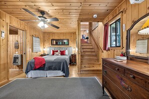 L’intérieur d’une cabane en bois, avec un lit, une commode et un escalier menant à un étage supérieur.
