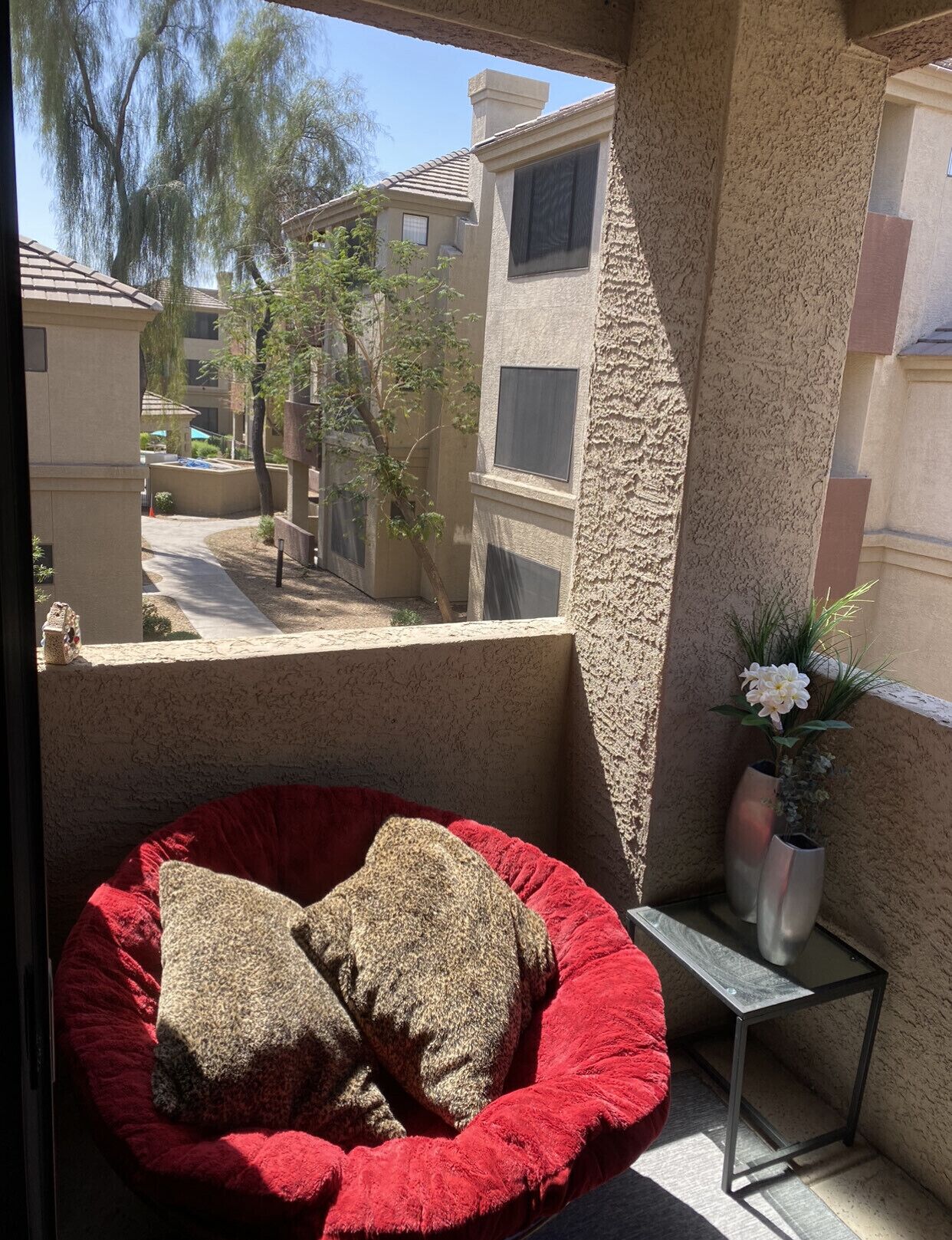 A balcony with a red bean bag chair and two pillows, a small metal table, and a potted plant.