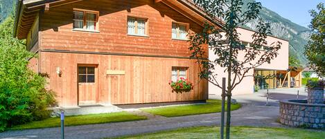 A wooden chalet with a sloped roof, a front entrance, and a small garden area with a tree and flower bed.