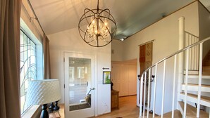 Een ruime kamer met een kroonluchter, een houten bureau met een lamp, een koeienhuidtapijt en een trap.
