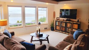Een woonkamer met een groot raam met uitzicht op een wateroppervlak, een aan de muur bevestigde televisie en een houten plafond.