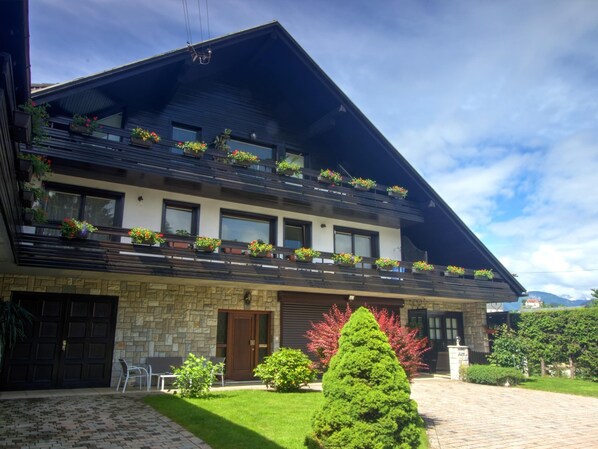 A two-story building with a stone facade, multiple balconies with flower boxes, and a paved driveway.
