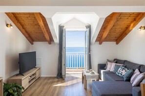A cozy living room with a TV, a sofa, and a balcony with a view of the sea.