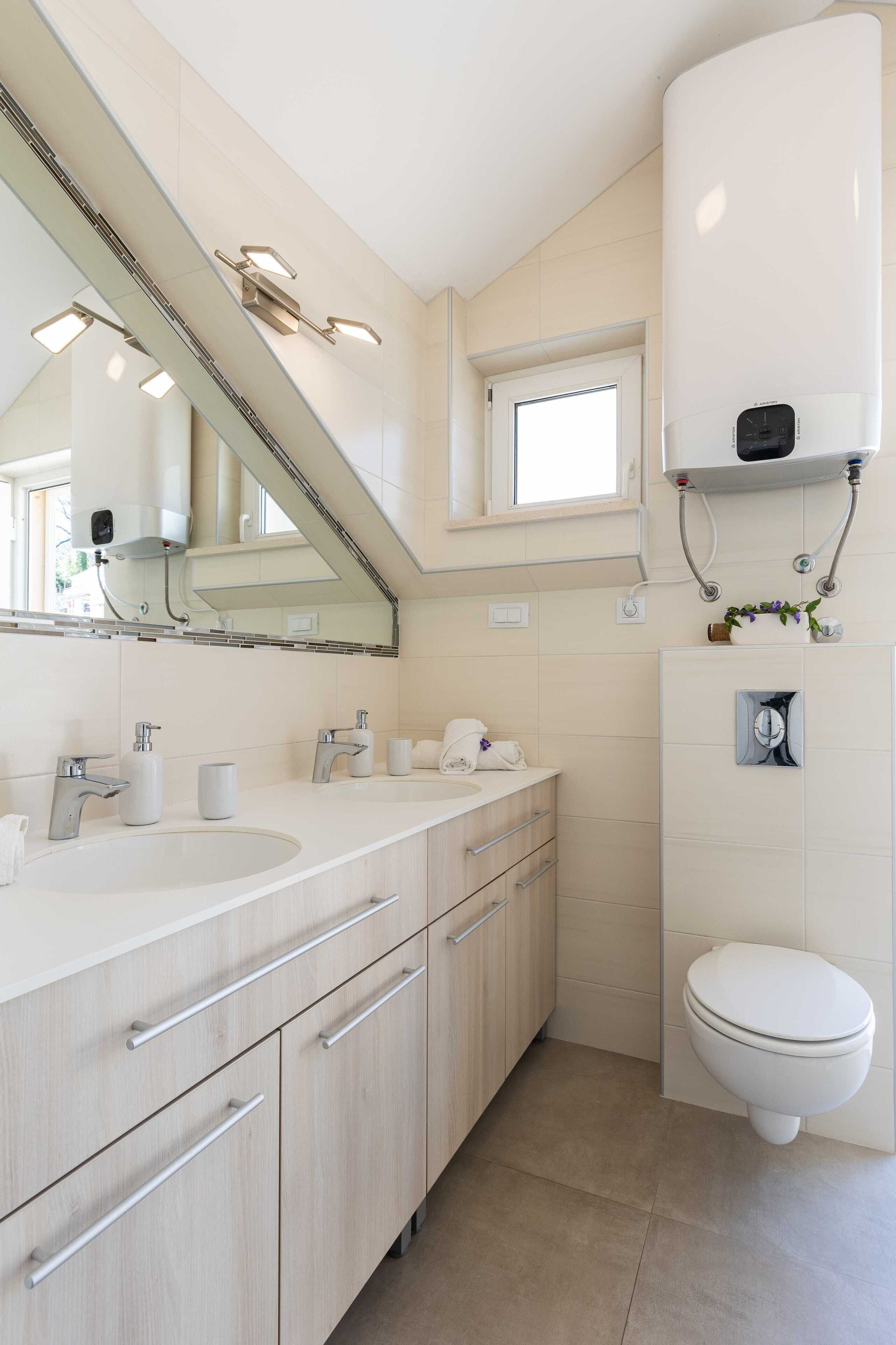 A modern bathroom with a large mirror, two sinks, a toilet, and a window above the toilet.
