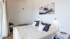 A bedroom with a large bed, bedside tables, a bench, a wall-mounted artwork, and a potted plant. - Apartment M&K (Mlini)