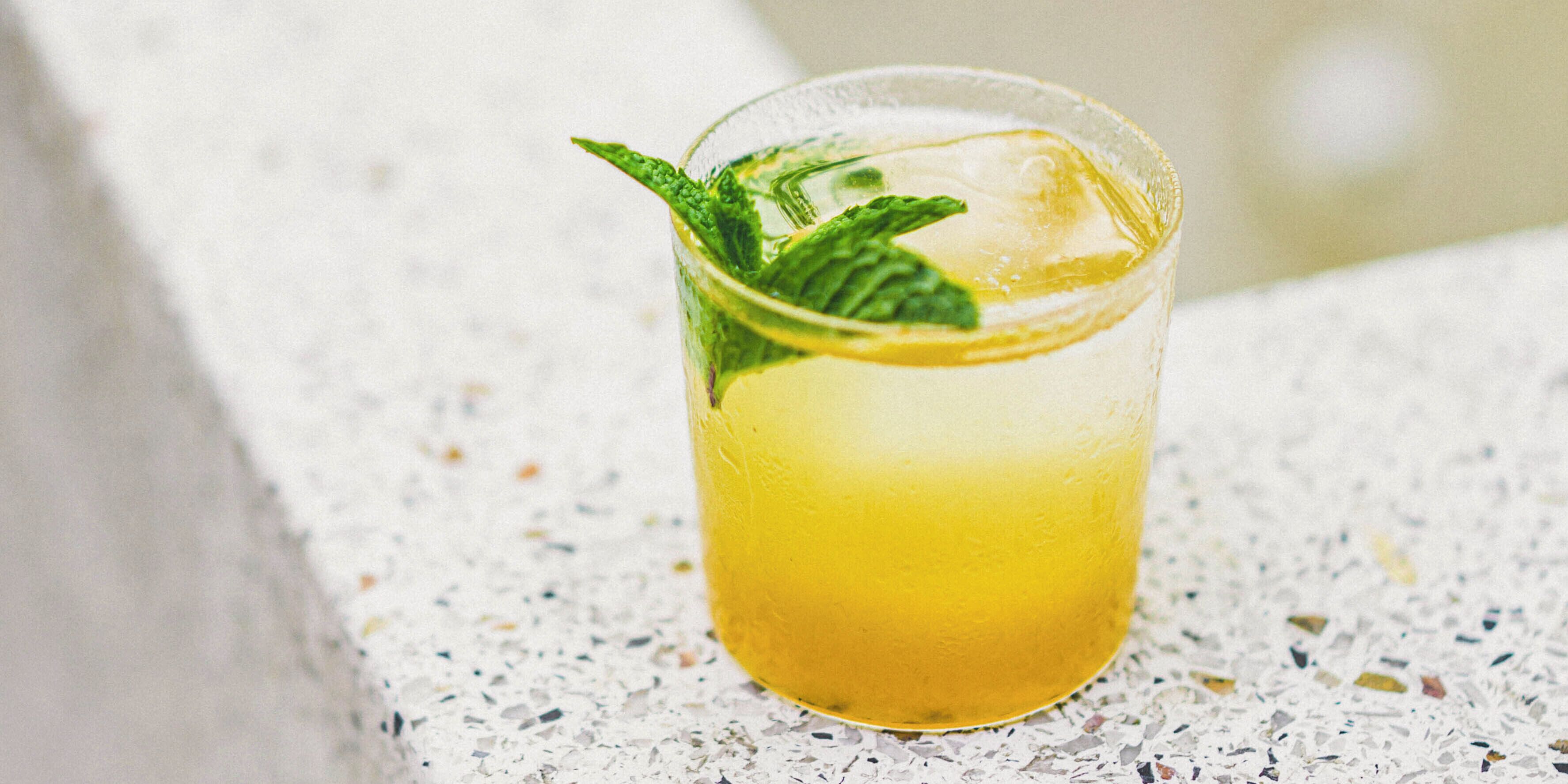 A glass of yellow beverage with mint leaves on a speckled surface.