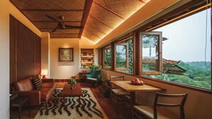 A cozy living room with a sofa, coffee table, and dining table, large windows with a view of greenery, and a ceiling fan. - The Sayan House (Ubud)