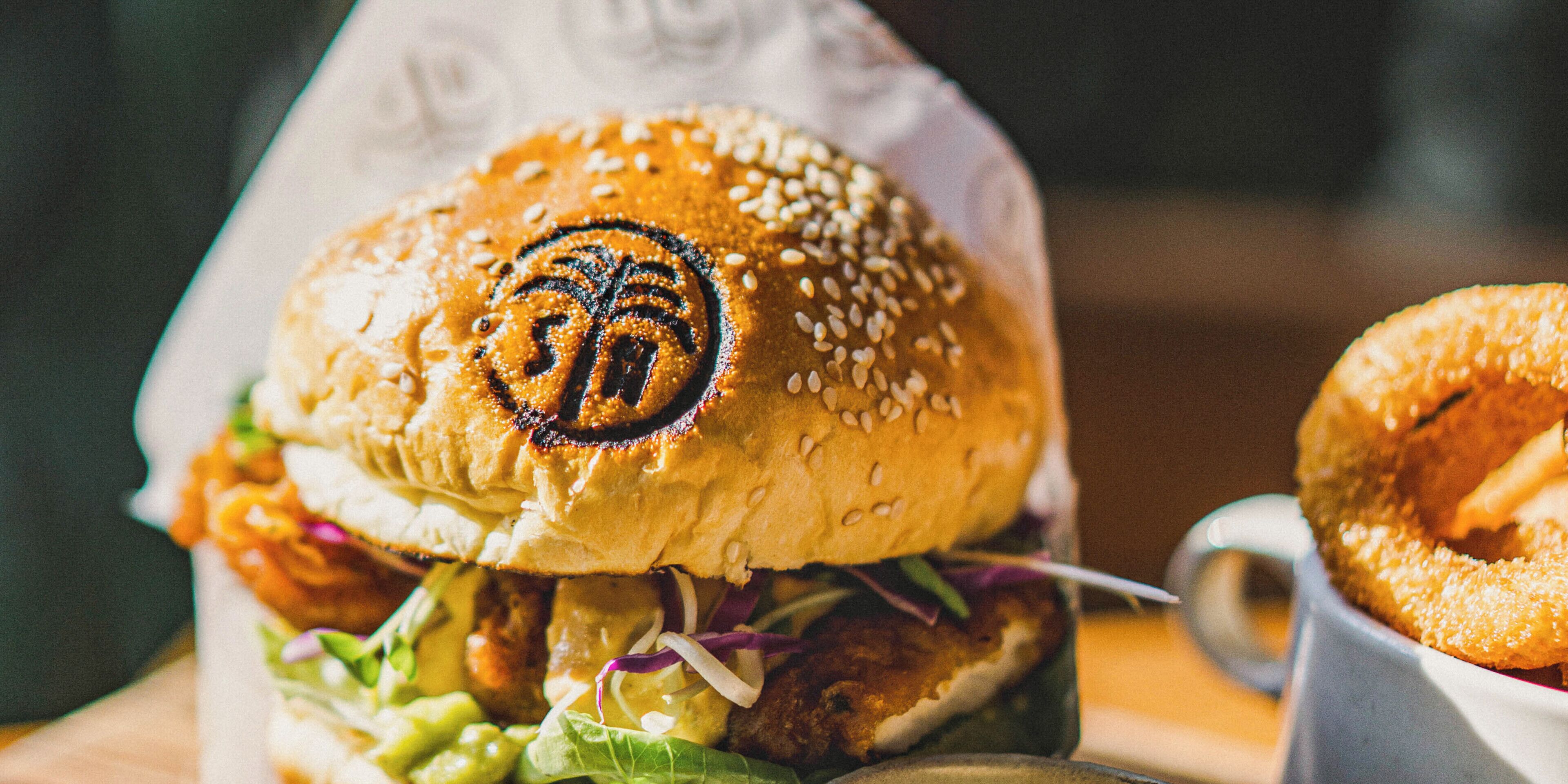 A burger with sesame seeds and a palm logo on the bun, accompanied by a side of onion rings.