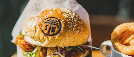 A burger with sesame seeds and a palm logo on the bun, accompanied by a side of onion rings.