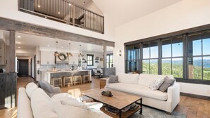 A spacious living area with a high ceiling, a large window offering a scenic view, and a kitchen with a central island. - Amazing Views @ Luxury Home, Outdoor Fireplace + Game Room (Grand Lake)