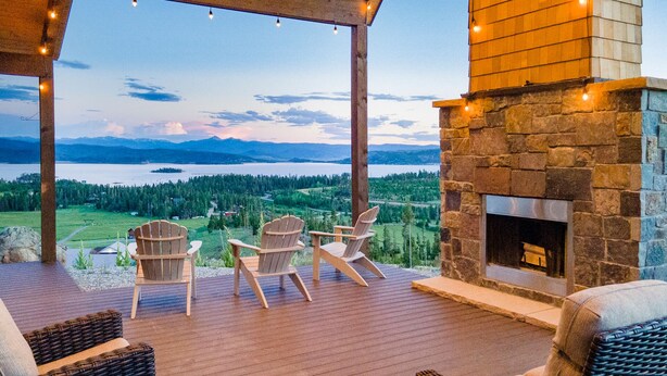 A covered outdoor seating area with Adirondack chairs, a stone fireplace, and a view of a lake and mountains. - Amazing Views @ Luxury Home, Outdoor Fireplace + Game Room (Grand Lake)