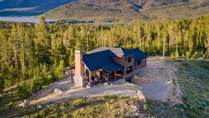 A modern two-story house with a large deck and a chimney, surrounded by dense forest and mountains. - Amazing Views @ Luxury Home, Outdoor Fireplace + Game Room (Grand Lake)