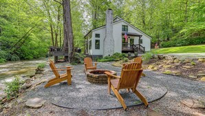 Una cabaña con chimenea de piedra, rodeada de sillas Adirondack, ubicada en un área boscosa con un río al fondo.