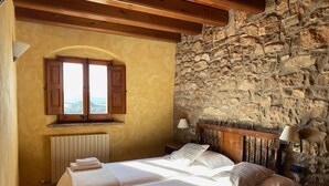 Un dormitorio con dos camas, una pared de piedra, vigas de madera, una ventana con contraventanas y un radiador.