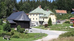 Un edificio tradizionale in legno con tetto spiovente, un edificio più grande giallo con tetto verde, e un muro in pietra sullo sfondo.