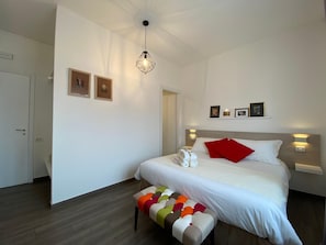 A modern hotel room with a neatly made bed, a colorful ottoman, and a pendant light.