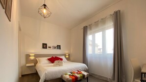 A modern hotel room with a neatly made bed, a bench with colorful cushions, a hanging light fixture, and a view of the city through large windows. - Economy Double Room Standard Rate (Cefalù)