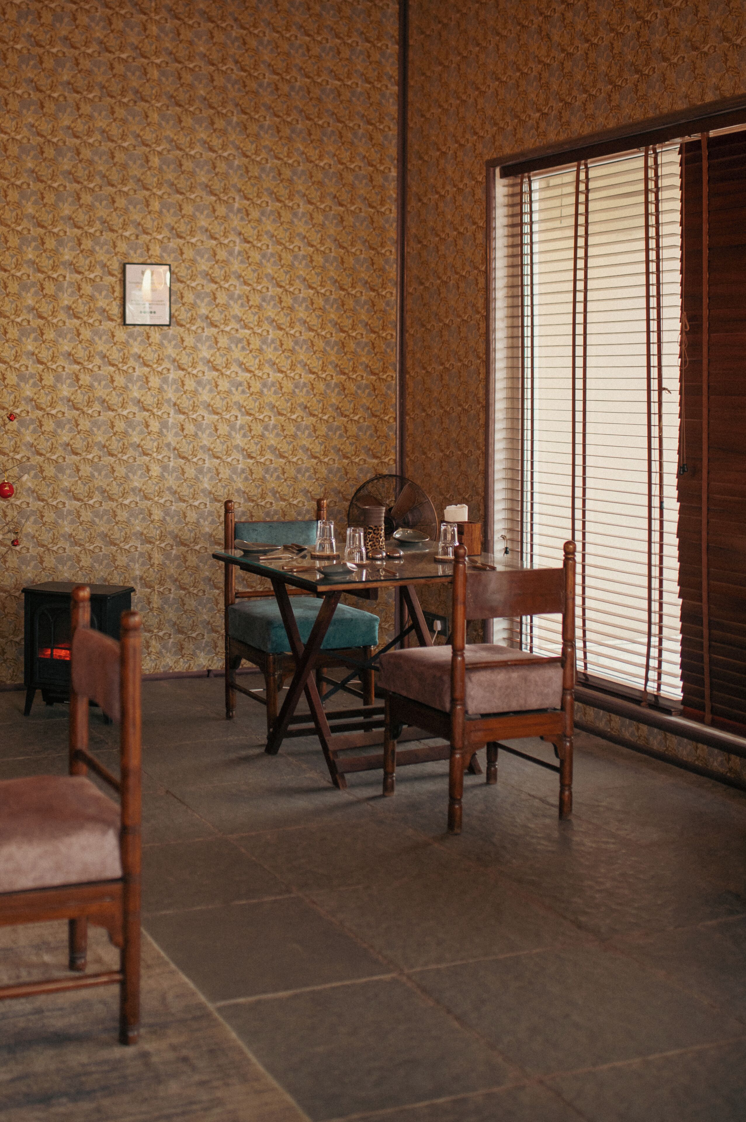 Un comedor con una mesa dispuesta para dos personas, un papel tapiz estampado y un amplio ventanal con persianas.