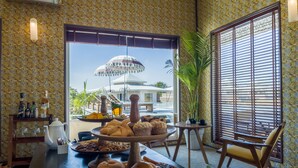A dining area with a variety of pastries and drinks on a table, a potted plant, and a view of a pool and building outside. - Brij Pola, Jawai - Luxury Jungle Camp with Private Pools (Bali)