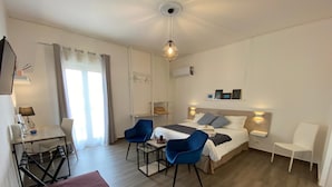 A modern hotel room with a bed, two blue chairs, a small table, a desk, and a red ottoman.