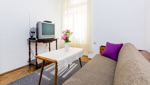 Una sala de estar con una televisión sobre una mesa de madera, un sofá con un cojín morado y un jarrón con flores.