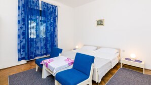 Un dormitorio con dos camas, una silla, una mesa con mantel floral, una alfombra pequeña y un cuadro en la pared.
