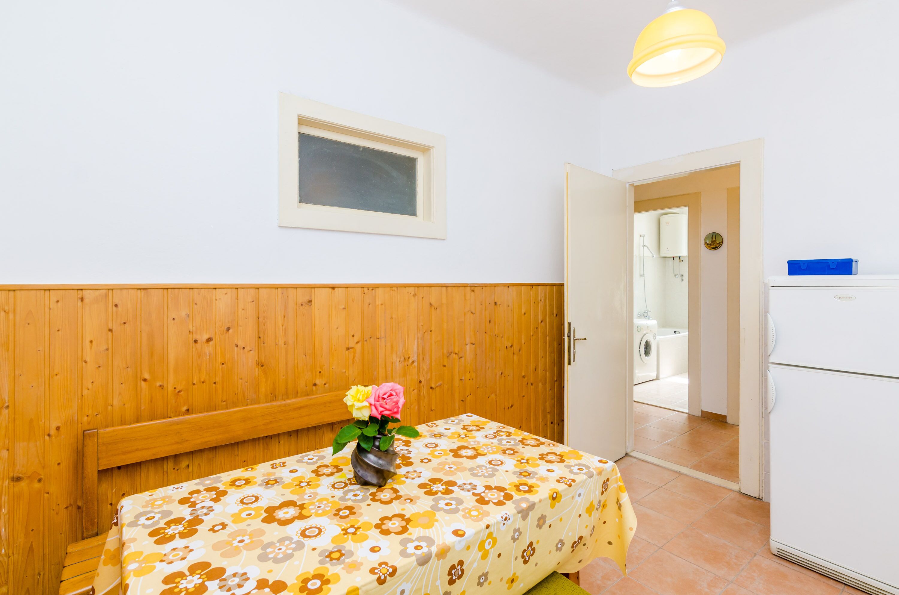 Une pièce avec un mur en bois, un lit recouvert d’une courtepointe à motifs floraux, une petite table sur laquelle est posé un vase de fleurs, une fenêtre et une porte donnant sur une salle de bain équipée d’un lave-linge.