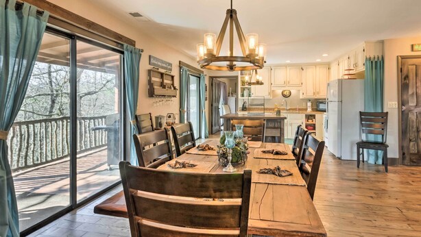 A dining area with a wooden table, chairs, and a chandelier. There is a kitchen in the background with cabinets and a refrigerator. - 'Joshua’s Lodge' w/ Hot Tub + Mountain Views! (Franklin)