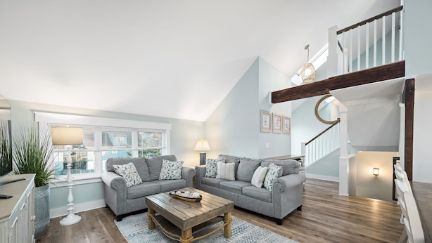 Un vaste espace de vie aménagé dans un style industriel, avec un plafond haut, un escalier en bois et une vue sur l’extérieur grâce à de grandes fenêtres.