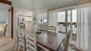 Un coin repas avec une table en bois et des chaises blanches, une cuisine en arrière-plan et un balcon agrémenté de rideaux blancs.