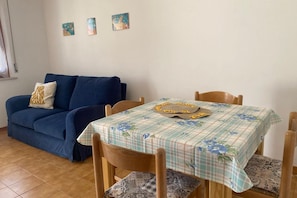 A living room with a blue sofa, a dining table with a floral tablecloth, and three wall-mounted artworks.