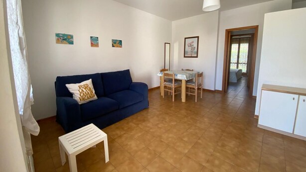 A living room with a blue sofa, a small white table, a dining table with chairs, and a kitchen area with cabinets.