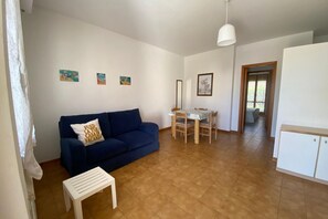 A living room with a blue sofa, a small white table, a dining table with chairs, and a kitchen area with cabinets.