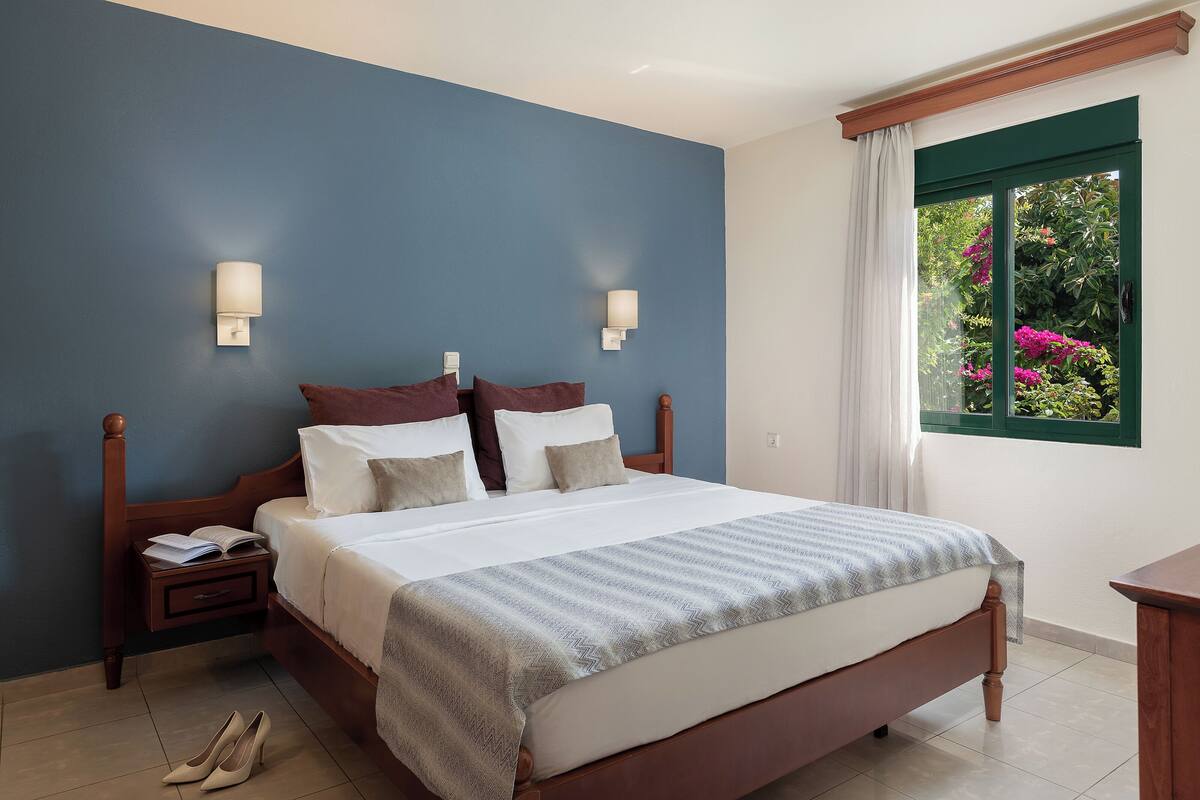 A bedroom with a large bed, two bedside tables, a nightstand, and a window with a view of greenery.