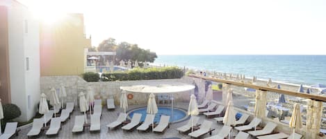 A pool area with sun loungers and umbrellas, a view of the sea, and a stone wall.