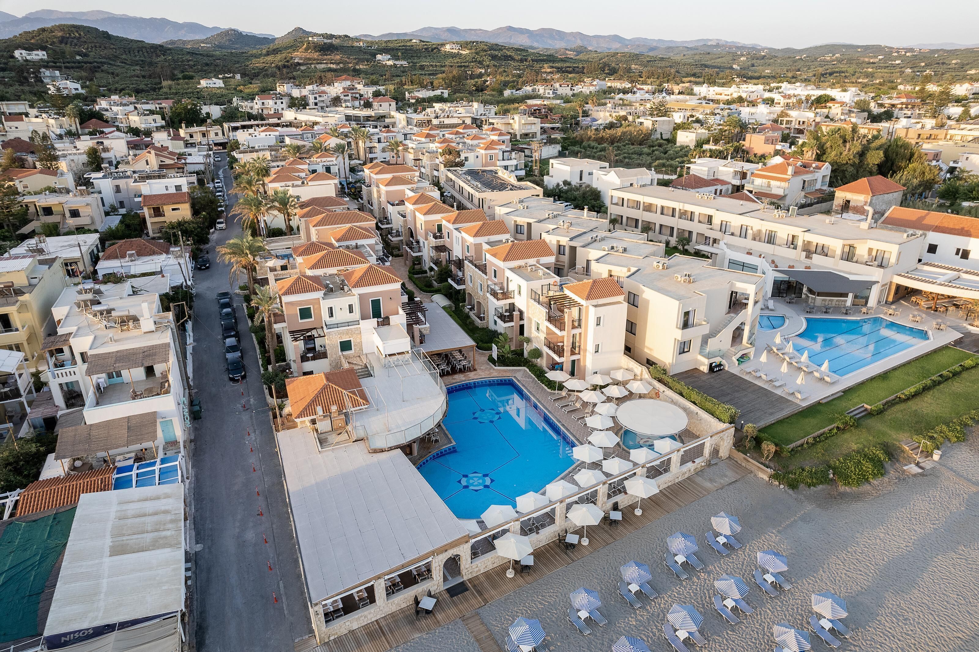 A resort with multiple buildings, a central swimming pool, and a beach area with sun loungers.