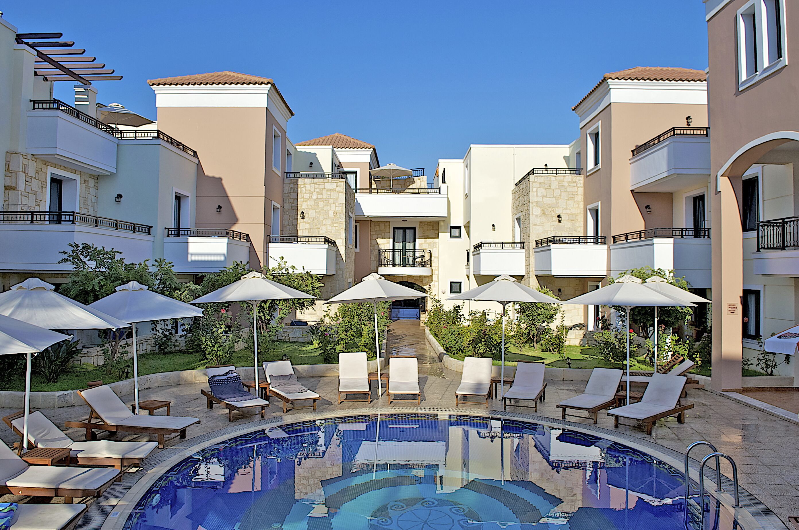 A pool area with lounge chairs and umbrellas, surrounded by buildings with balconies and a clear sky.
