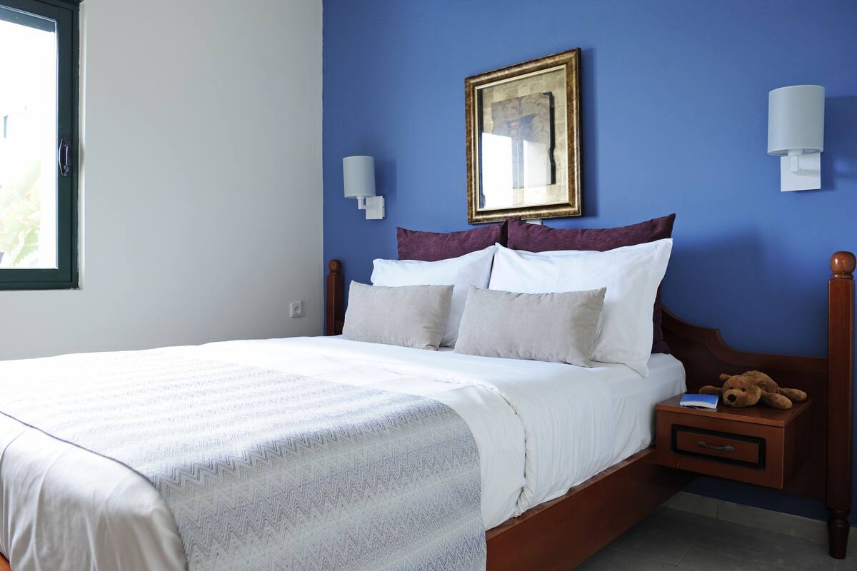 A neatly made bed with white linens and a striped blanket, a wooden nightstand with a teddy bear, and a framed picture on the wall.