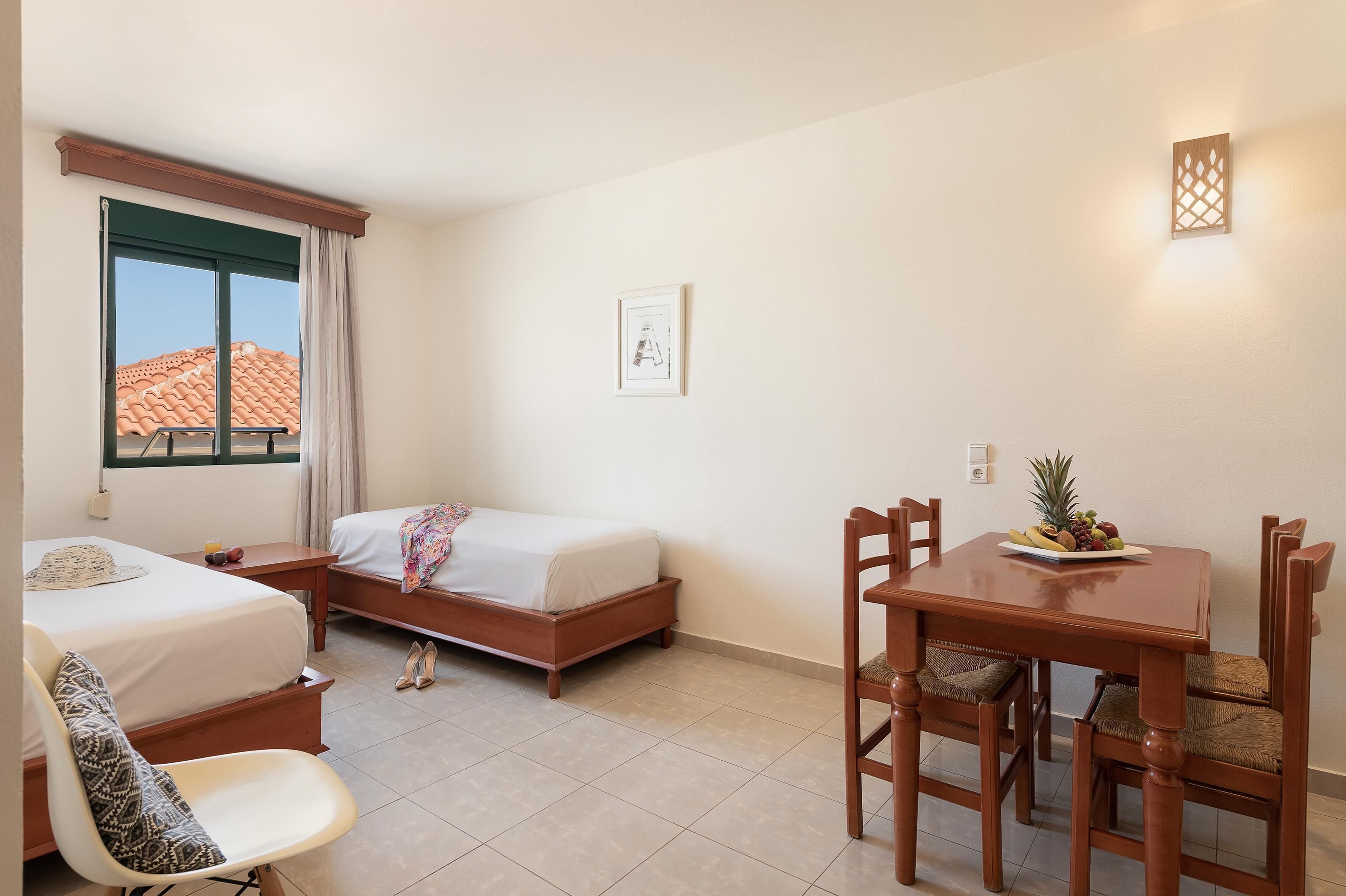 Ein Hotelzimmer mit zwei Betten, einem Esstisch mit Stühlen, einem kleinen Tisch mit Obstschale, einem Bild an der Wand und einem Fenster mit Vorhängen.