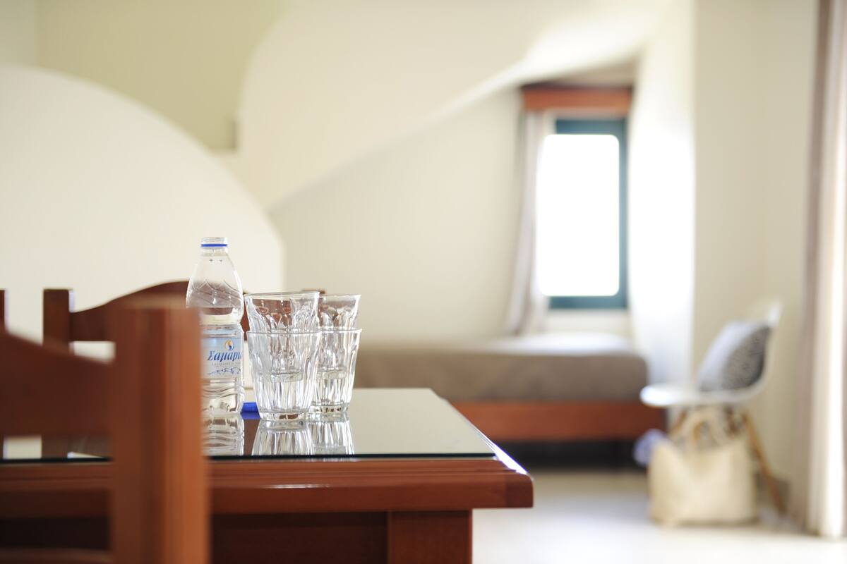 A table with a water bottle and glasses, a chair, and a bag in a room with a window and a sofa.