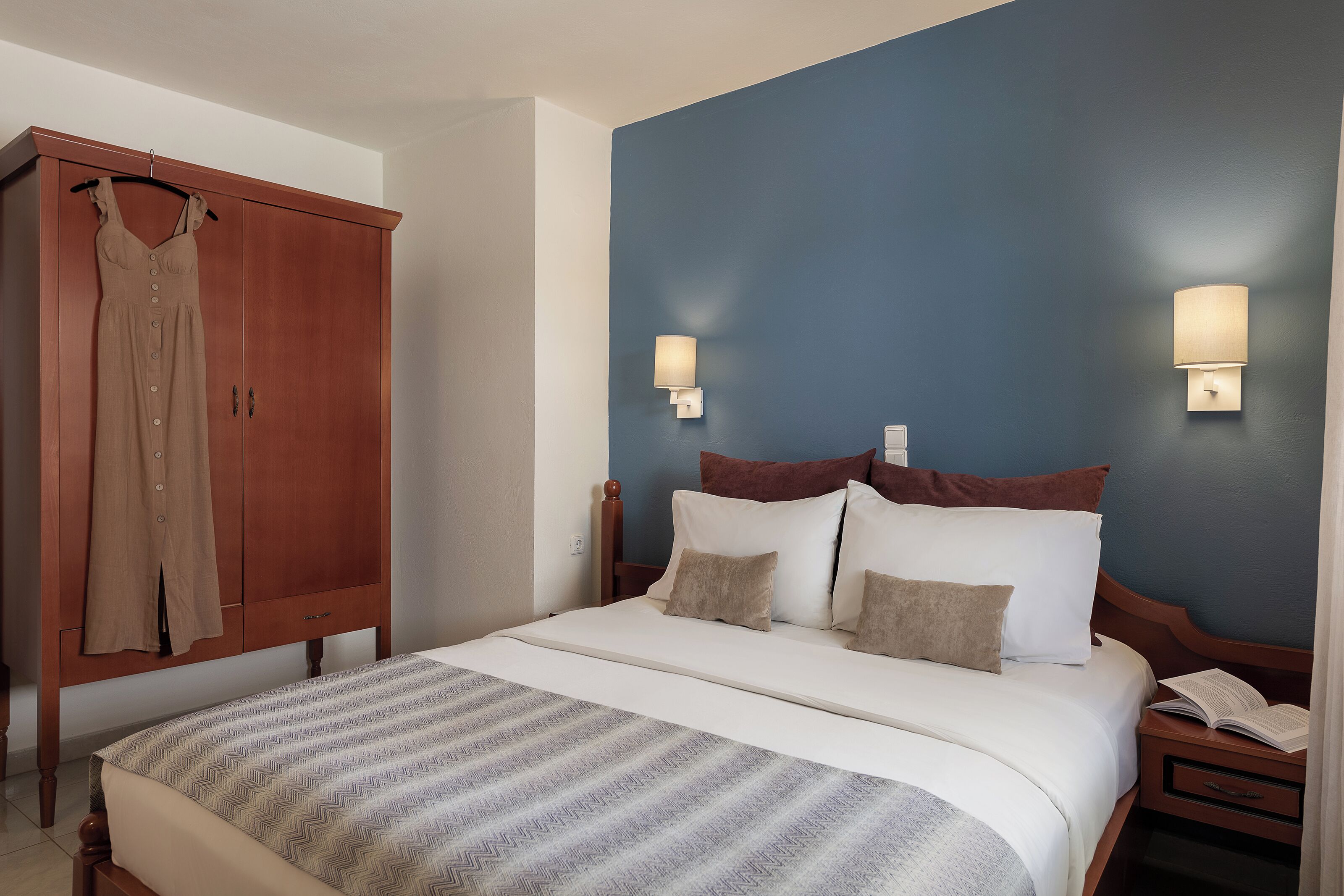 Une chambre à coucher comprenant un lit, une armoire, deux lampes fixées au mur et une table de chevet avec un livre.