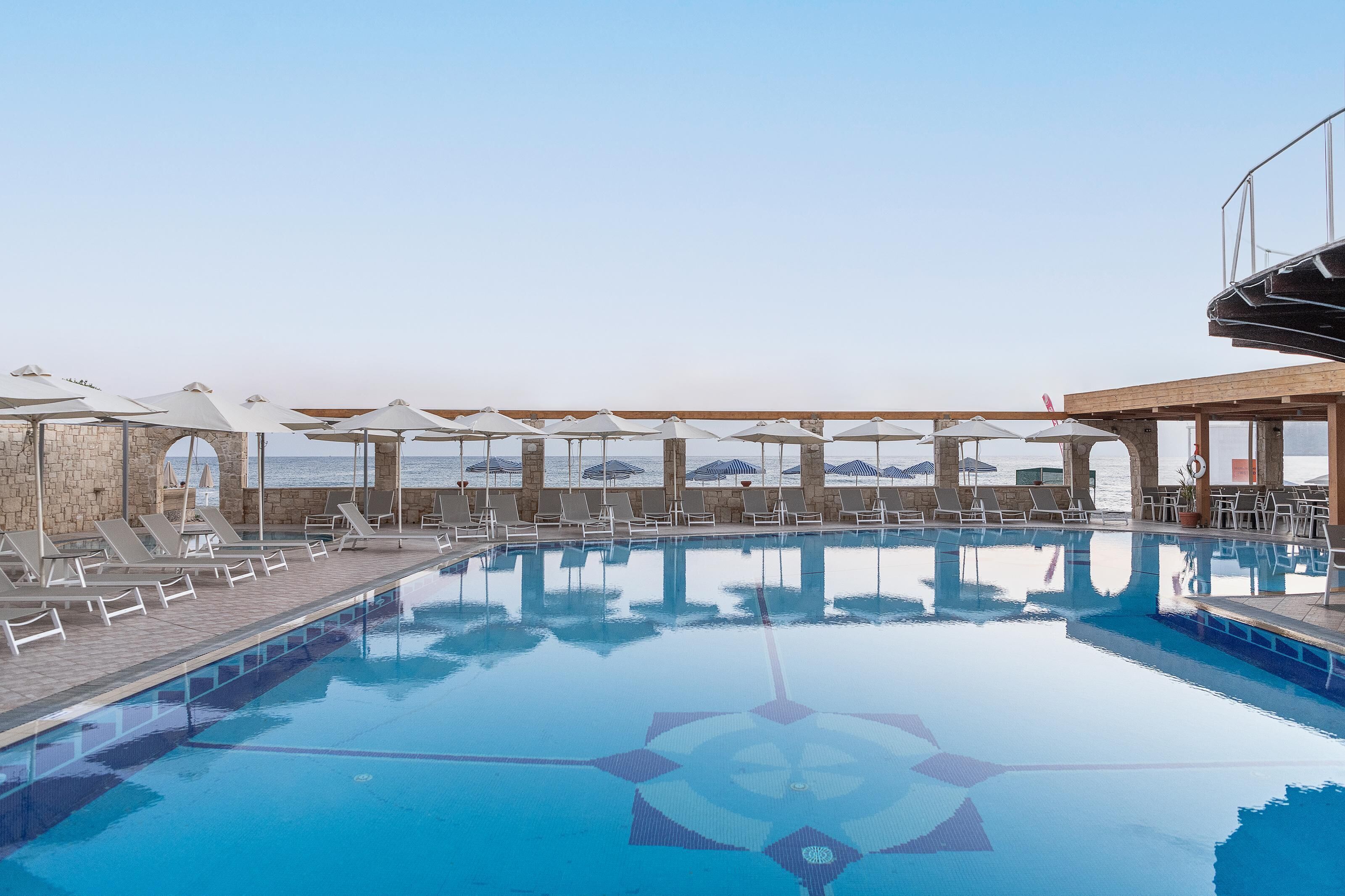 A pool area with lounge chairs and umbrellas, a stone wall, and a view of the sea.