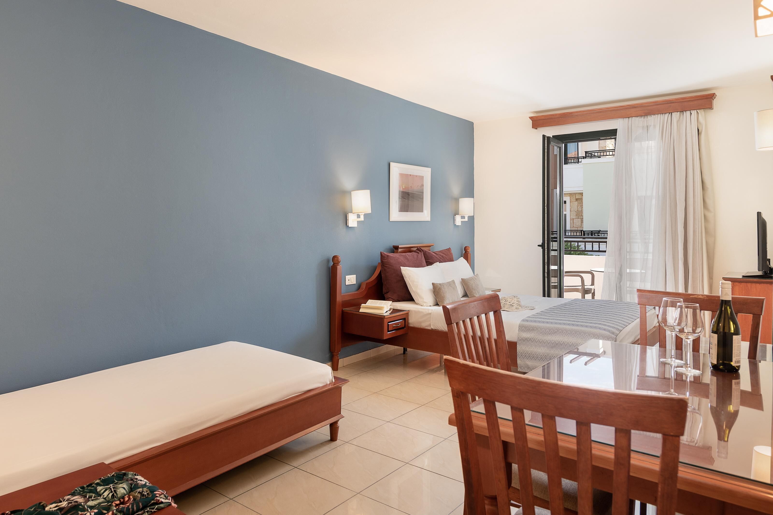 A hotel room with a bed, a small table with chairs, a bedside table with a lamp, and a view of the outside balcony.