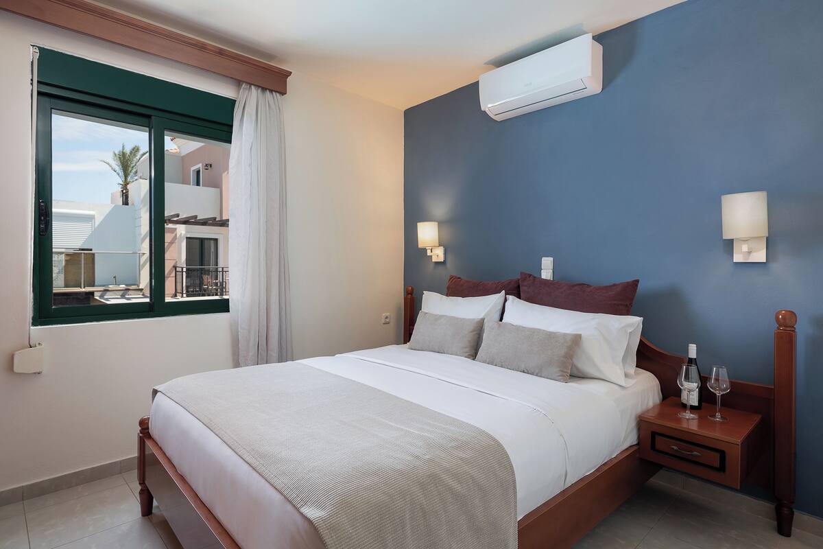 A neatly made bed with a wooden headboard, a bedside table with a bottle and glass, and an air conditioning unit on the wall.