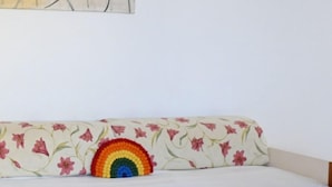 Ein Zimmer mit einem Bett, einem blumigen Kissen, einem regenbogenfarbenen Gegenstand auf dem Kissen und abstrakter Wandkunst.