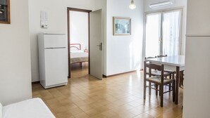 A compact kitchen with a refrigerator, a dining table with chairs, a bed in the adjacent room, and a window with curtains. - M271 - Marcelli, comfortable two-room apartment with balcony (Marcelli)