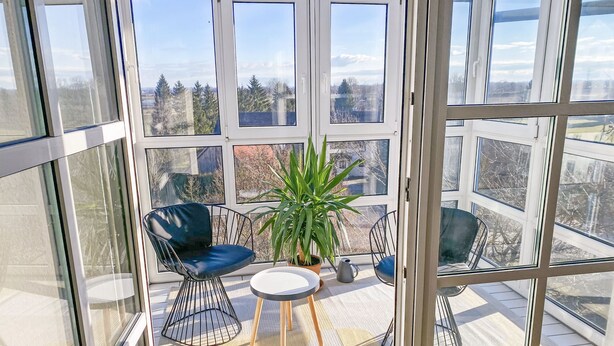 Une véranda avec un toit en verre, deux chaises, une petite table et une plante en pot.